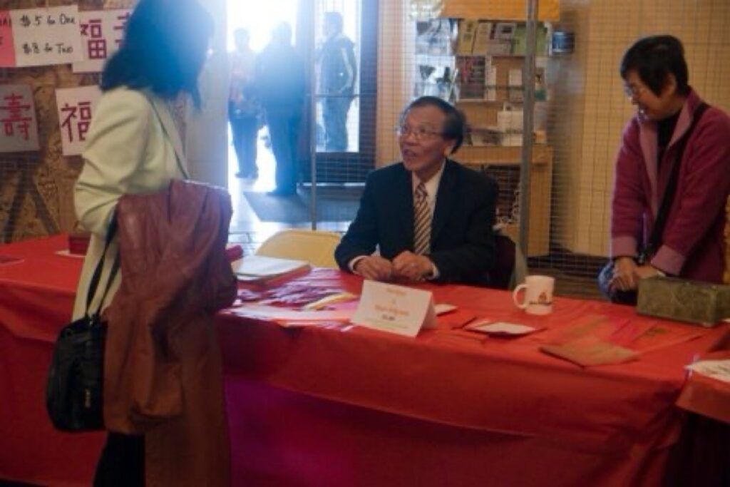 Dr. Henry Wong sitting behind a red table listening to his wife Sylvia, who is in front of the table, inside the Bob Hope Theater in Stockton.