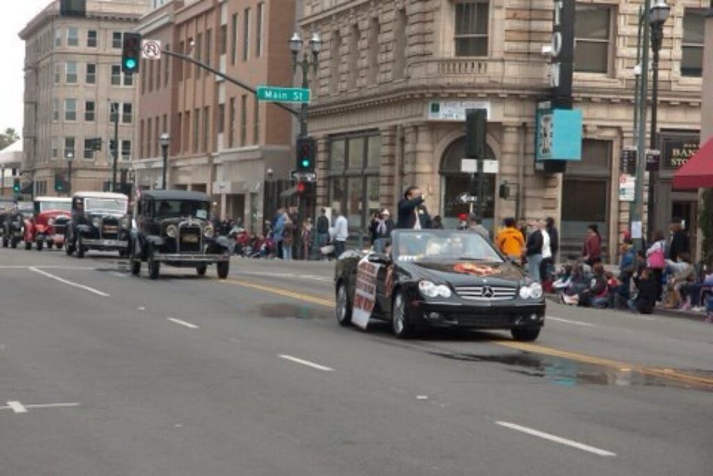 Dr. Henry Wong waves to the crowd while standing in the back of a black Mercedes-Benz convertible as it drives in the middle of the road in Downtown Stockton. Several cars from the 1920s or 1930s follows.