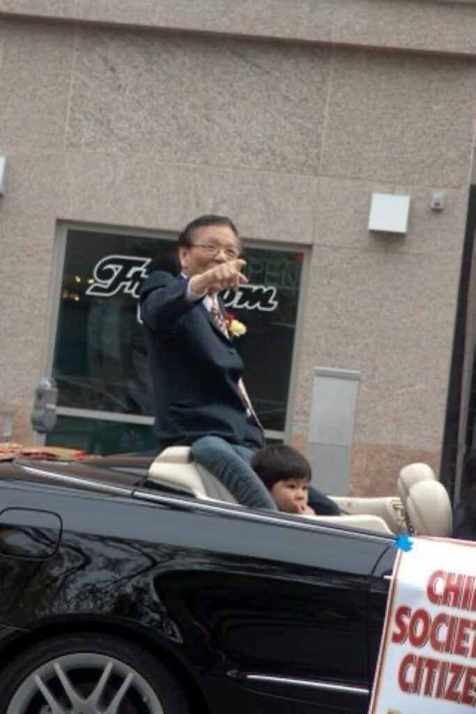 Dr. Wong points with his right index finger while sitting on the rear deck of a top-down, black Mercedes-Benz convertible. His grandson is sitting next to him in a baby seat.