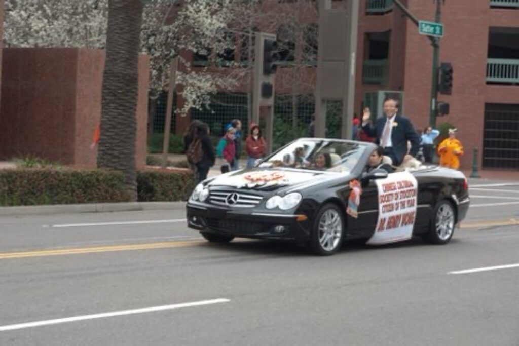 Dr. Henry Wong waves while standing in the rear of a top-down, black Mercedes-Benz Convertible. His son is driving and his wife Sylvia is in the passenger seat.