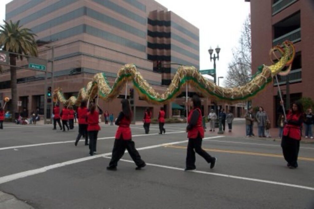 Volunteers wearing red vests, black long-sleeved shirts, and black pants holding up a long gold and green dragon with metal sticks march down the street in Stockton's Chinese New Year parade.