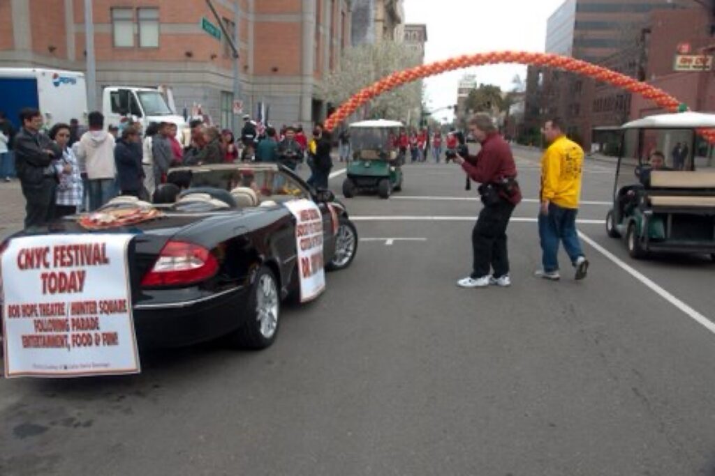 A photographer wearing a red shirt takes a photo of the black Mercedes-Benz convertible. A white sign is attached to the back of the convertible that reads Civic Festival Today.