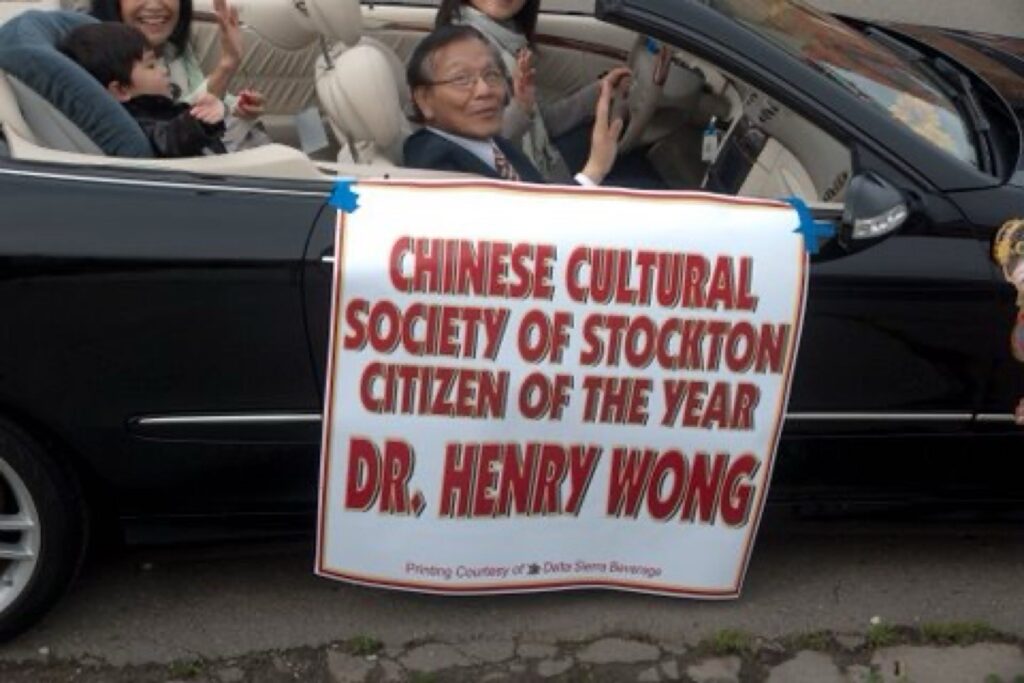 Dr. Henry Wong waves from the passenger seat of the black Mercedes-Benz convertible. His wife Sylvia is also waving from the rear seat next to their grandson sitting in a baby seat.