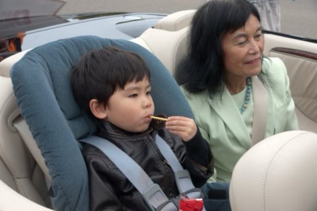 A young boy sitting in a baby seat and sucking on a popsicle is sitting next to his grandmother Sylvia Wong, who is wearing a light green dress, in the rear of a Mercedes-Benz convertible.