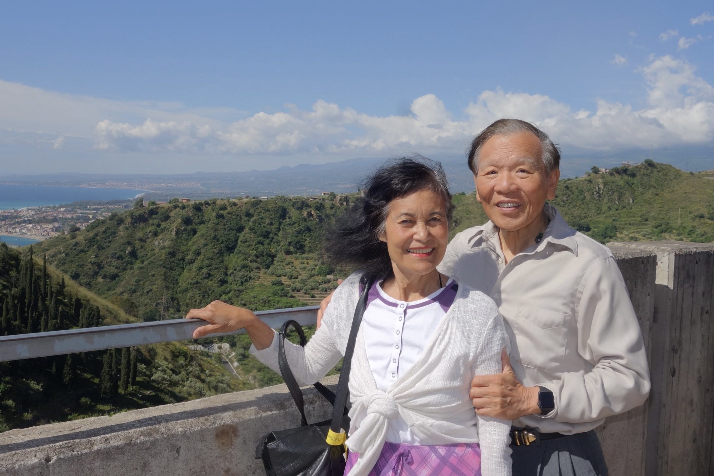 Sylvia and Henry Wong smiling while standing together in front of the Sicilian coast.