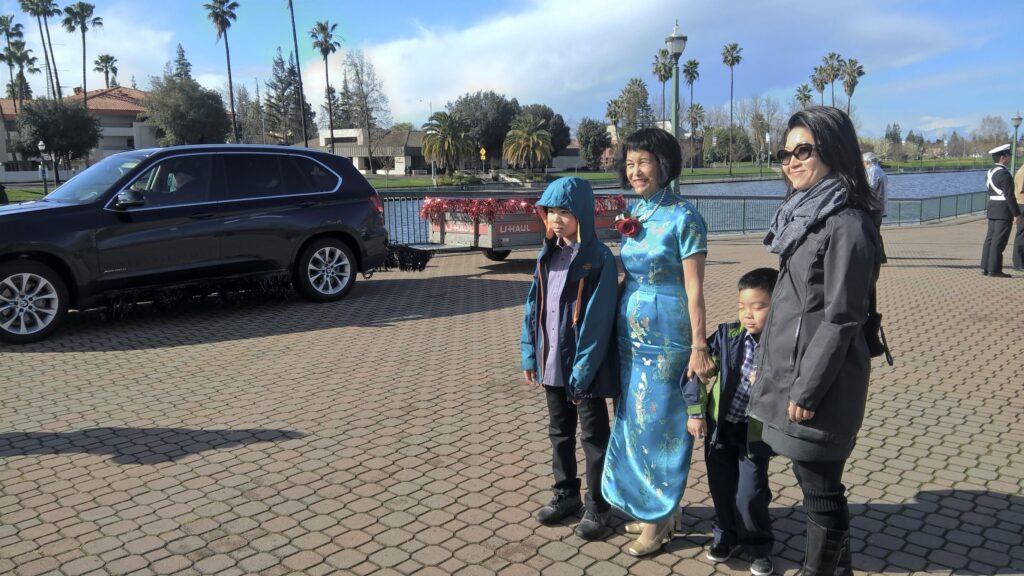 Sylvia Wong is wearing a long, turquoise, Chinese-style dress while standing next to her two grandsons and daughter-in-law.