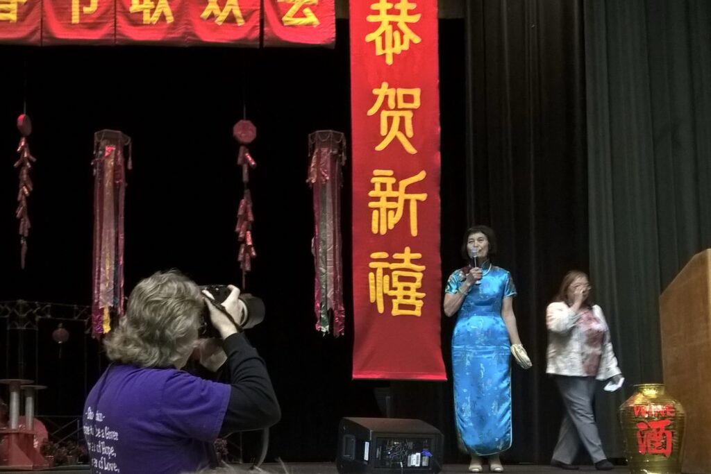 Sylvia Wong wearing a long shiny turquoise Chinese-style dress speaking to the crowd on stage inside the Stockton Civic Memorial Center. She is standing next tow red banners with Chinese characters.