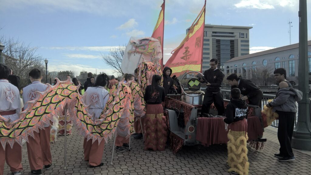 Volunteers from the Concord Kung Fu Academy prepare to enter the Chinese New Year Parade. They are holding a long pink, green, and black dragon and wearing white kung fu tops with orange pants.