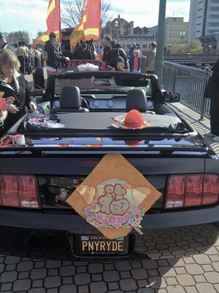 The black Ford Mustang Convertible (probably from 2007) that Sylvia rode in had a fabric sign taped to the back with a single Chinese character and a California license plate that read PNYRYDE.