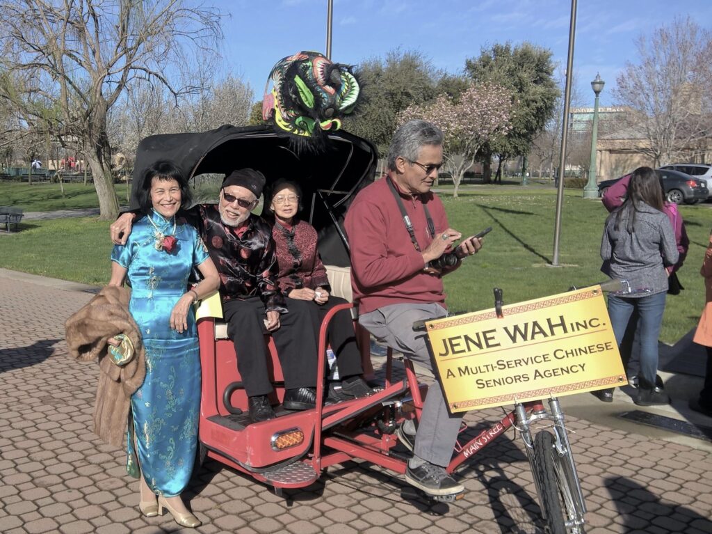 Sylvia Wong sits next to a red human-powered carriage that have two people sitting inside it. The driver is looking at a phone. A sign on the front of the carriage reads "Jene Wah Inc.: A Multi-Service chinese Seniors Agency."